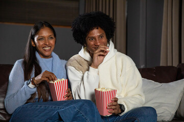 Diverse couple sitting on dark sofa in home living room eating from popcorn tubs © wavebreak3