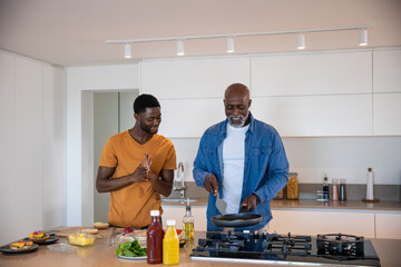 African american father and son cooking breakfast in home kitchen with spatula, frying pan