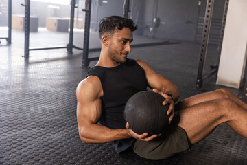 Man in 20s holding medicine ball, rotating torso and exercising on gym floor with squat racks