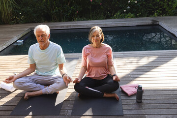 Senior couple wearing activewear meditating on black yoga mats on poolside deck with water bottle