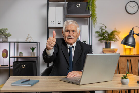 Like. Happy Caucasian elderly businessman using laptop looking approvingly at camera showing thumbs up sign positive something good great news positive feedback. Grandpa sitting at home office table.