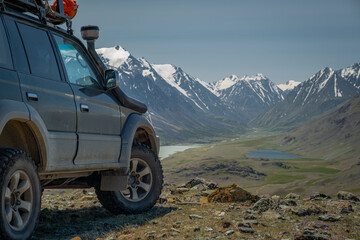 A dark off-road vehicle with aggressive mud tires and a snorkel is parked on a rocky mountain pass, overlooking a wide green valley, lake, and snow-capped peaks under a blue sky during a summer advent © ANDREY