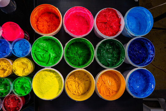 Colorful Ink for Screen Printing FactoriesInside a busy Tee shirt factory plastisol inks in multiple tones bright and dark are neatly arranged for industrial use. Workers use handmade tools handles