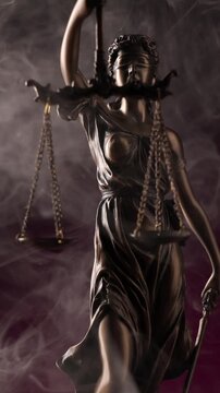 Dramatic white smoke flowing around the bronze scales and blindfolded face of a Lady Justice statue set against a deep purple background.