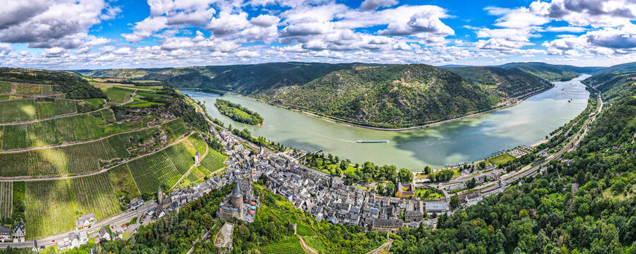 Panoramic aerial view of the Rhine River valley with vineyards, historic town, castles and winding river surrounded by green hills under a dramatic sky.