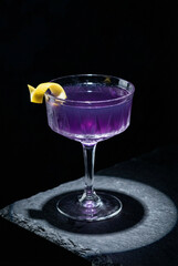 An elegant purple Aviation cocktail in a classic coupe glass, garnished with a lemon twist, on a slate coaster against a dark, moody background.