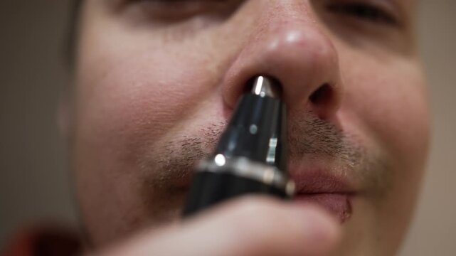 Close-up shot of a man utilizing a compact electric trimmer to eradicate excess hair from his nostril. His attention to grooming exemplifies a typical aspect of a man's daily self-care regimen