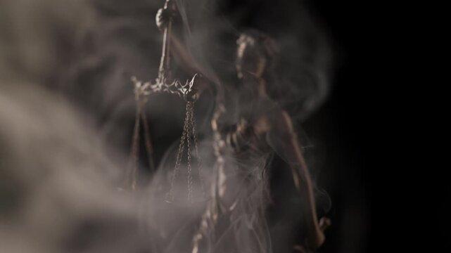 Dense grey mist drifts across the frame, slowly dissipating to unveil the bronze figure of a blindfolded Lady Justice holding balanced scales in a dramatic studio setting.