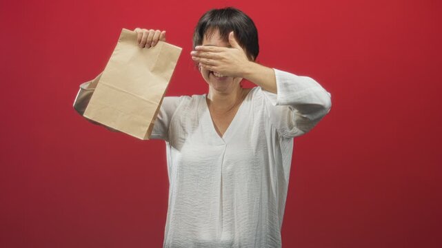 Woman holds paper bag with hand covering eyes, smiling and peeking in a red studio; playful shyness.