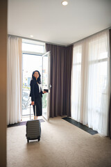 Smiling woman with travel suitcase and documents standing at window
