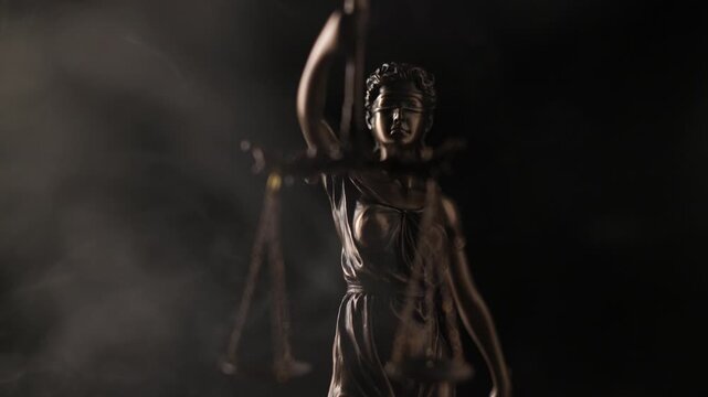 Wisps of white smoke drift slowly around the illuminated bronze figure of a blindfolded Lady Justice statue holding scales against a dark background.