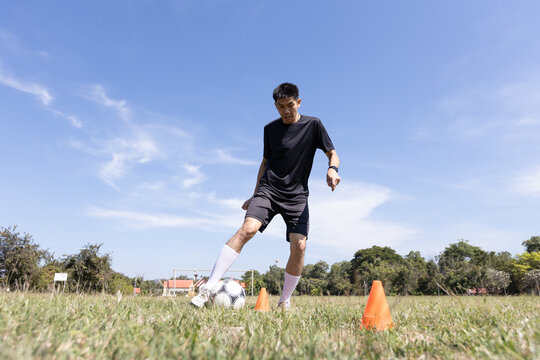 An adult Asian man practices soccer dribbling with cones and a football on a grass field outdoors. Suitable for concepts related to sports training, fitness, active lifestyle, and football.