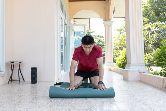 An adult Asian man kneels on a yoga mat while preparing it for exercise on a home terrace during daytime. Suitable for concepts related to home workout, fitness routine, active lifestyle, wellness.