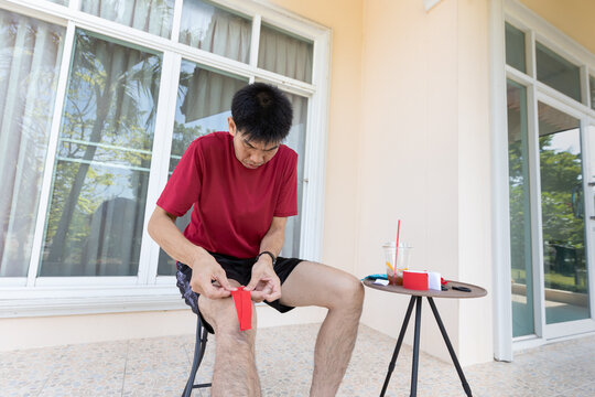 An adult Asian man applies kinesiology tape on his knee while sitting at home during daytime. Suitable for concepts related to sports preparation, fitness routine, muscle support, and active lifestyle