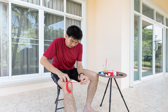An adult Asian man applies kinesiology tape on his knee while sitting at home during daytime. Suitable for concepts related to sports preparation, fitness routine, muscle support, and active lifestyle