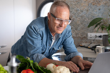Smiling mature man searching for new recipes on his laptop while cooking healthy food at home in a...