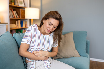 Woman suffering from stomach ache or abdominal pain sitting on sofa at home, female feeling menstrual cramps or digestive problems and holding belly, health issues and food poisoning concept