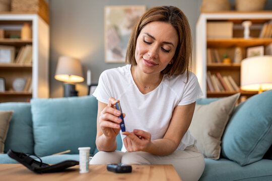 Young woman testing blood sugar levels with a glucose meter and lancet device at home managing diabetes or hypoglycemia sitting on sofa with medical equipment and testing strips