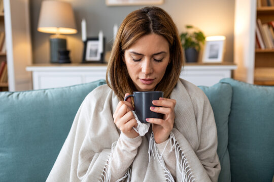 Sick young woman wrapped in a blanket sitting on the sofa and drinking a cup of hot tea to soothe a sore throat or cold symptoms at home during flu recovery and medical treatment concept