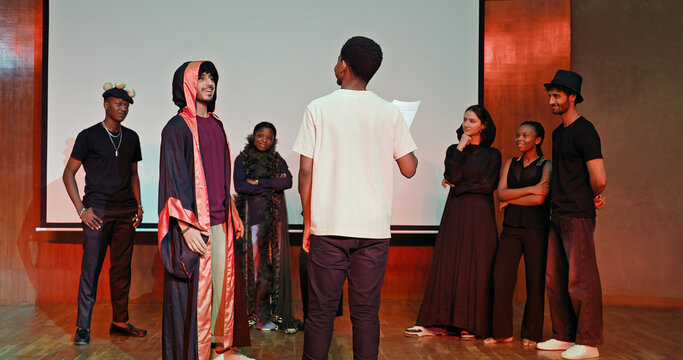 African young adult gen z male man hold scripts paper read play story lines scene standing indoor stage hall. Happy group boy girl enjoy do work show drama practice day inside campus place