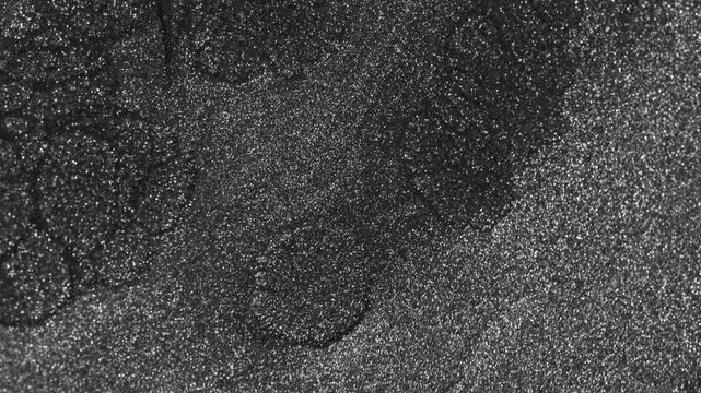 Intense shot of dark titanium paint separating and expanding into large cellular shapes while revealing a sandy glittering texture that spreads outwards on black background