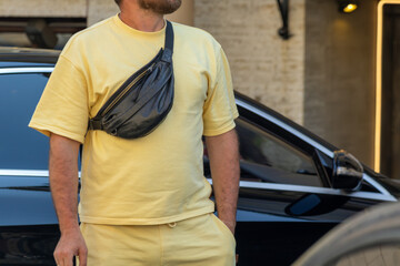 Man in casual clothing stands near parked car with crossbody waist bag across chest. Building entrance and wall lamp visible behind.