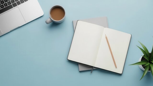 Overhead view of open notebook with pencil, coffee mug, laptop and plant on blue desk table workspace