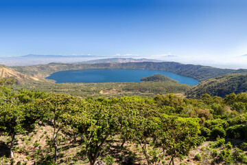 Lake Coatepeque in El Salvador, a deep blue volcanic crater lake and popular tourist destination near Santa Ana, surrounded by forested hills beneath a clear blue sky.