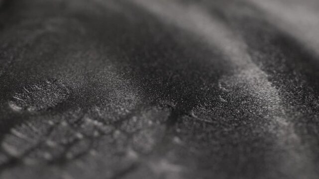 Rack focus shot of dark metallic steel paint with specialized sandy texture shifting sharpness across heavy waves and ridges while sparkling radiant glitter particles catch light on black background