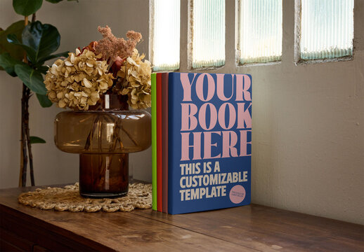 Mockup from real photo of four books placed on a wooden table next to a vase with dried flowers. 