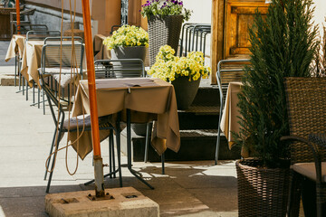 Outdoor cafe terrace shows empty tables with tablecloths beside steps and large flower planters. Doorway, wicker chair, and potted shrub visible near seating area.