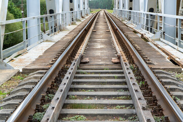 Fototapeta premium perspective capturing the transition from concrete railroad sleepers to the timber deck of a massive white steel truss bridge featuring parallel rusty iron rails