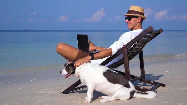 Relaxed young freelancer in a hat working on a laptop computer while sitting on a beach chair and petting his dog, enjoying a serene vacation on a beautiful tropical island with a turquoise sea