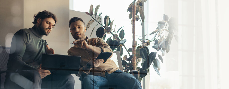 Two men are seated in a bright room, engaged in a work discussion. One man points to a laptop while the other takes notes. Plants grow nearby, adding to the environment.