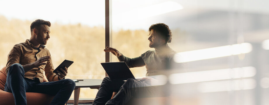 Two men are engaged in conversation in a bright office. One man holds a tablet while the other has a laptop in front of him. They appear to share thoughts and ideas.