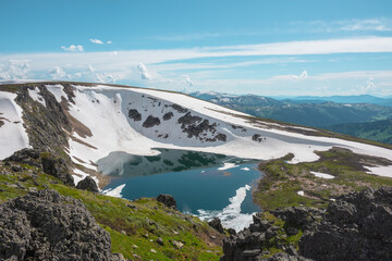 Scenic sunlit landscape with alpine lake in rocky snowy cirque near stone hill top during thaw. Ice floats in mountain lake among rocks and snows with view to forest mountain range under cloudy sky. © Daniil