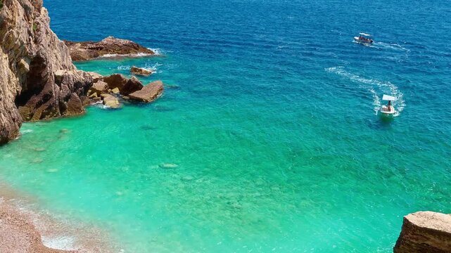 Aerial view of small motor boats cruising in the turquoise Adriatic Sea along a pebbled beach in Montenegro. A picturesque Mediterranean summer vacation scene with crystal clear water and cliffs.
