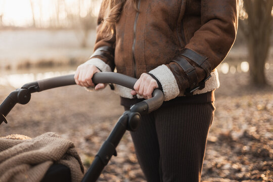 A person wearing a brown shearling jacket pushes a pram through a park at sunset. The image focuses on the hands on the handlebar with a soft, natural background.