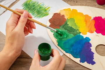 A woman holds a brush in her left hand and applies green paint to a palette. Creative mess on the table. Photo. Lifestyle.