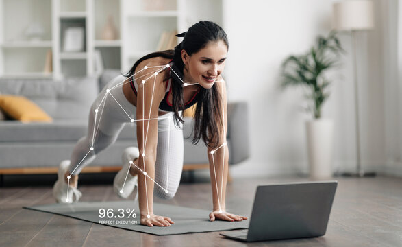 A woman performs a workout at home while watching an online fitness video. She tracks her performance with metrics displayed on the screen. The room has a simple design with a couch and plants. - Powered by Adobe