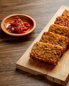 Fried tempeh slices arranged on wooden board with sambal tomat in small bowl, traditional Indonesian soybean snack photographed in minimal food styling 