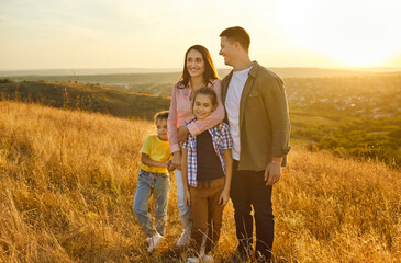 Obraz premium Happy family standing together at sunset, parents hug children. Father, mother, daughter and son enjoy golden light on meadow and hills in nature, calm moment of love and happiness of weekend hike