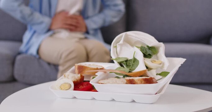 Sick man suffering from stomach pain and cramps, diarrhea and nausea after takeaway fast food eating at home. Person holding belly and sitting on sofa beside takeout box of leftovers meals on table