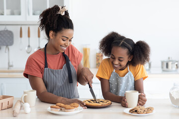 Excited black mom and daughter in aprons tasting homemade berry pie, cutting cake and smiling, kitchen interior. Happy african american mother and kid teen girl baking together at home, copy space © Prostock-studio