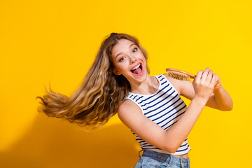 Energetic young woman smiling while brushing her long blonde hair, standing against a vibrant...