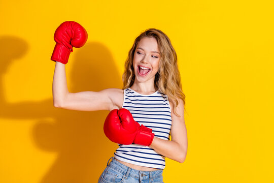 Happy young woman with boxing gloves flexing arm in a cheerful pose on a vibrant yellow background