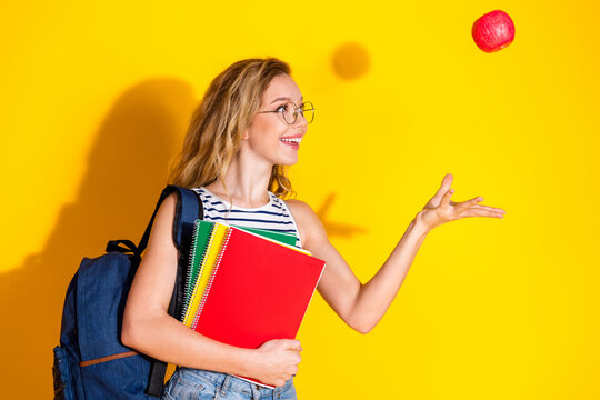 Cheerful young woman with blonde hair holding book and tossing an apple against a vibrant yellow background
