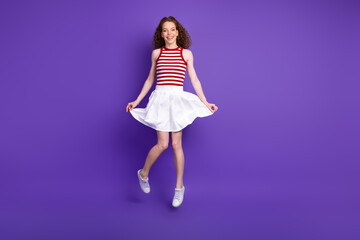 Naklejka premium Young woman in red and white striped top and white skirt jumps against purple background smiling and playful fashion moment