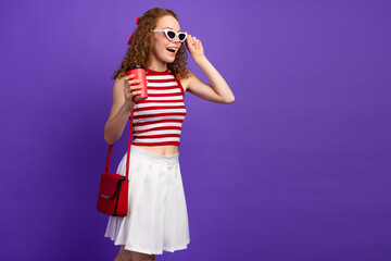 Stylish young woman with red curly hair in a striped top and white skirt holding a coffee cup...