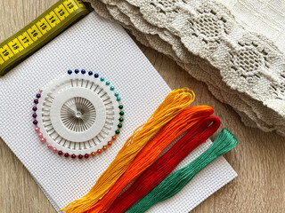 Multicolored floss threads, white canvas, sewing pins, and a centimeter ribbon lie on the table. Hobby or profession.